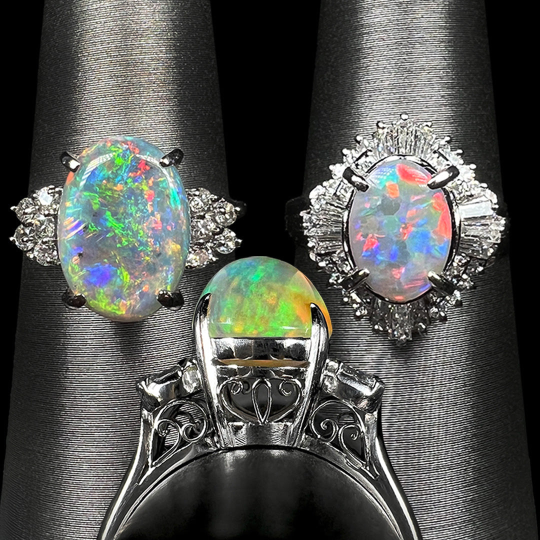 Three platinum opal and diamond rings shown on ring displays.  Two are showing facing the camera, and one is shown from its side gallery angle, showing filigree details.