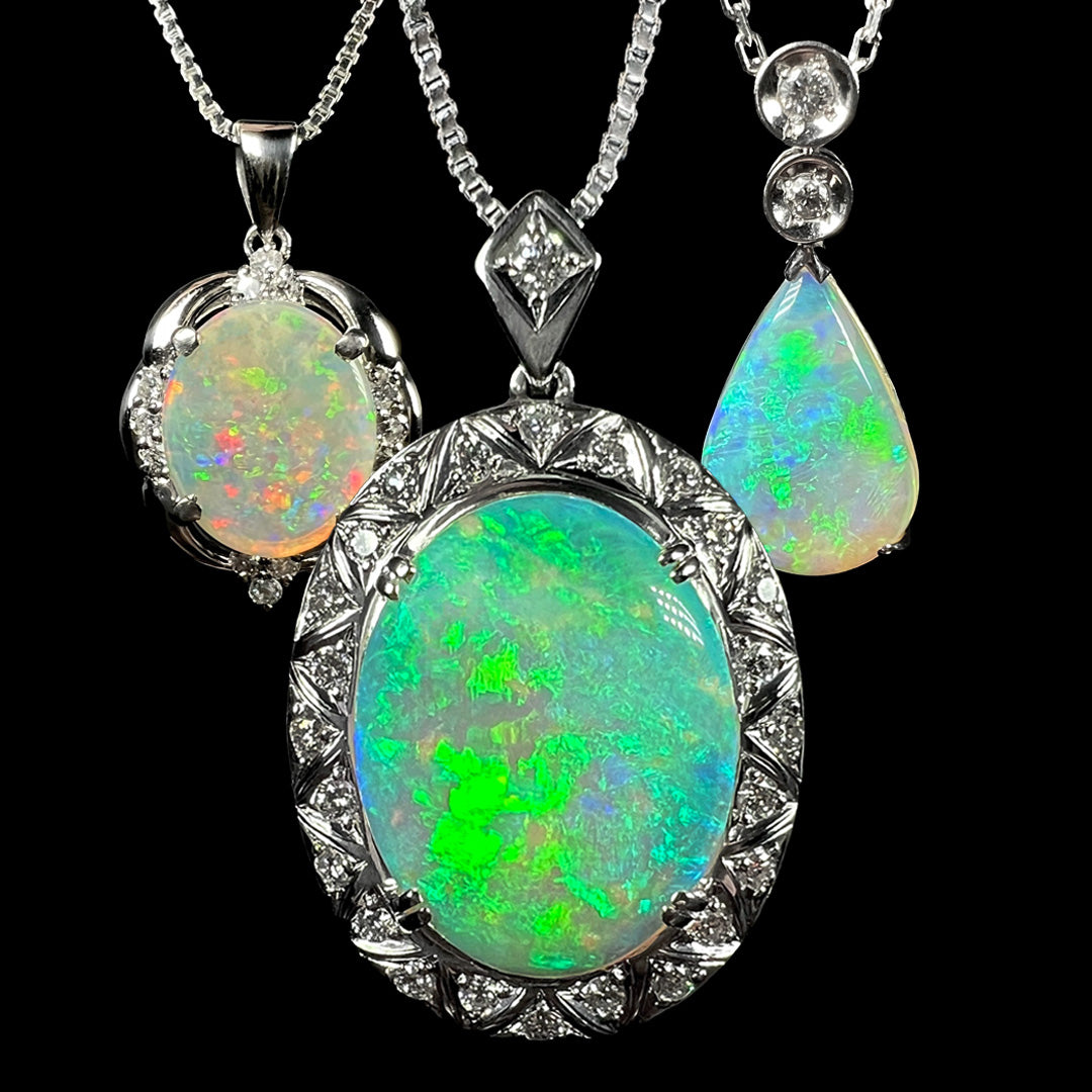 Three platinum Australian opal and diamond pendants shown hanging from chains against a black background.  Two opals are oval shaped, and one is pear shaped.