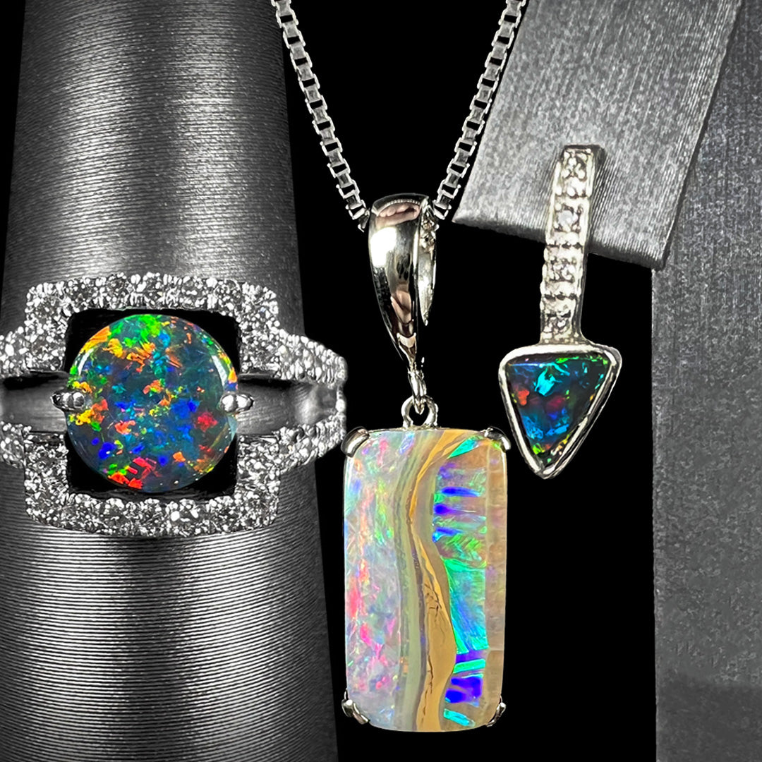 Three pieces of white gold opal jewelry; from left to right, a black opal and diamond ring, a rectangular boulder opal necklace, and a triangle shaped black opal drop earring.