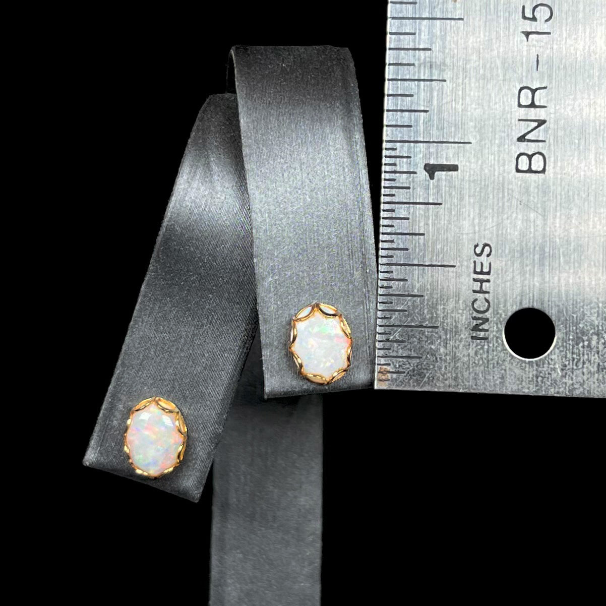 A pair of 0.25 inch-long Australian white opal stud earrings in yellow gold, shown on a ring display next to a ruler.