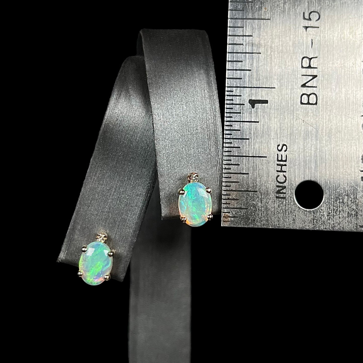 A pair of 0.33 inch-long Australian crystal opal and diamond stud earrings shown on an earring display next to a ruler.
