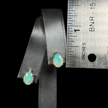 A pair of 0.33 inch-long Australian crystal opal and diamond stud earrings shown on an earring display next to a ruler.
