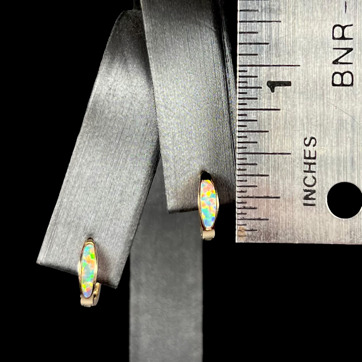 A pair of 0.33 inch-long yellow gold lab-created opal inlay huggie hoop earrings, shown on an earring display next to a ruler.