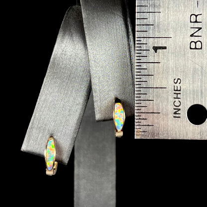 A pair of 0.33 inch-long yellow gold lab-created opal inlay huggie hoop earrings, shown on an earring display next to a ruler.