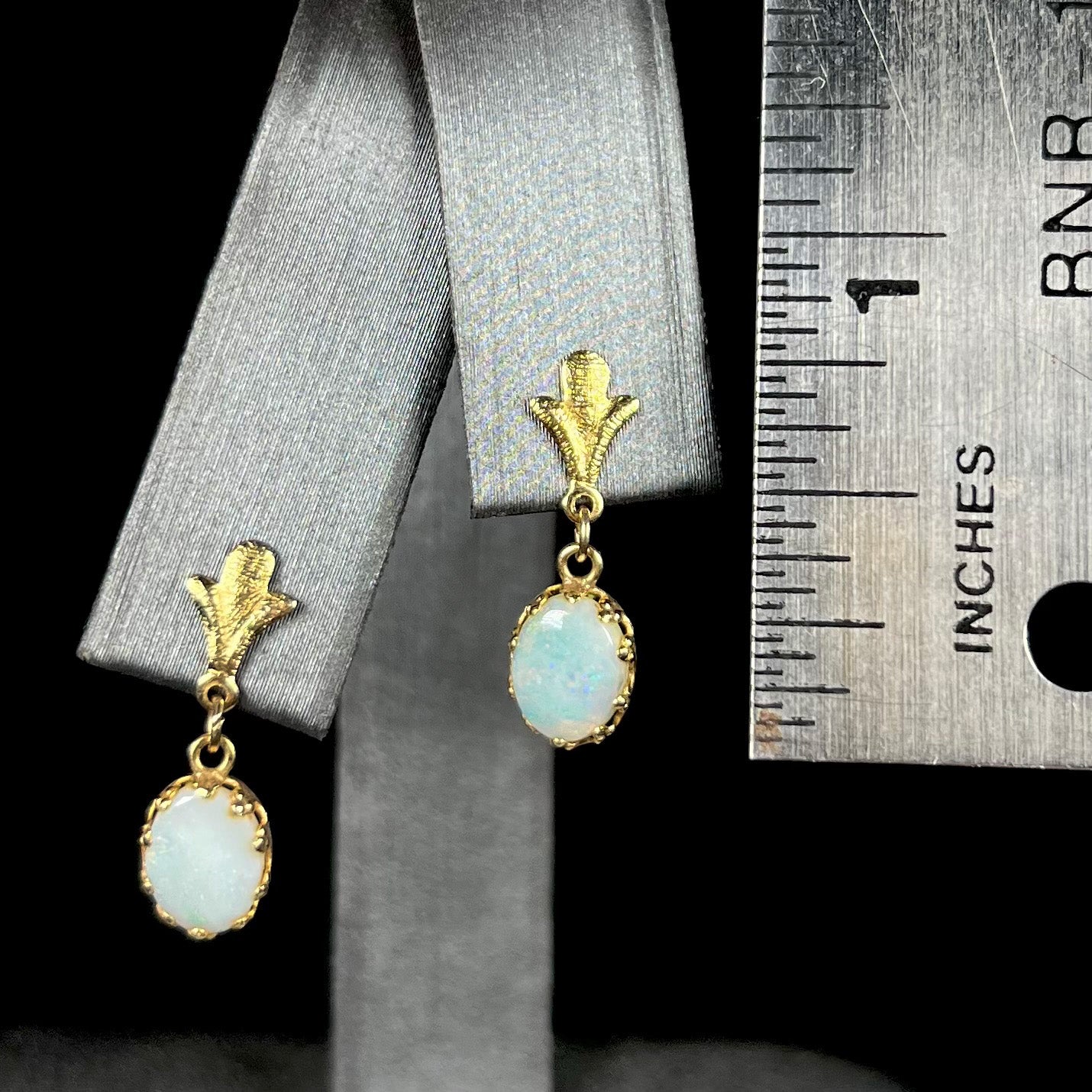 A pair of 0.75 inch-long yellow gold Fleur-de-Lis Australian light opal dangle earrings on an earring display next to a ruler.