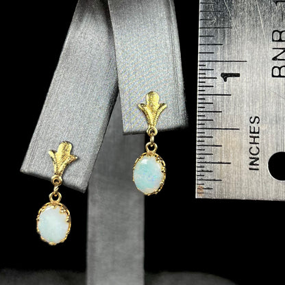 A pair of 0.75 inch-long yellow gold Fleur-de-Lis Australian light opal dangle earrings on an earring display next to a ruler.
