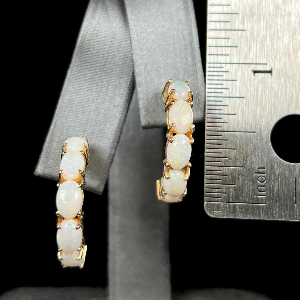Pair of large five-stone Australian white opal semi-hoop earrings in 14k gold measuring 0.9" long, shown on earring display next to ruler