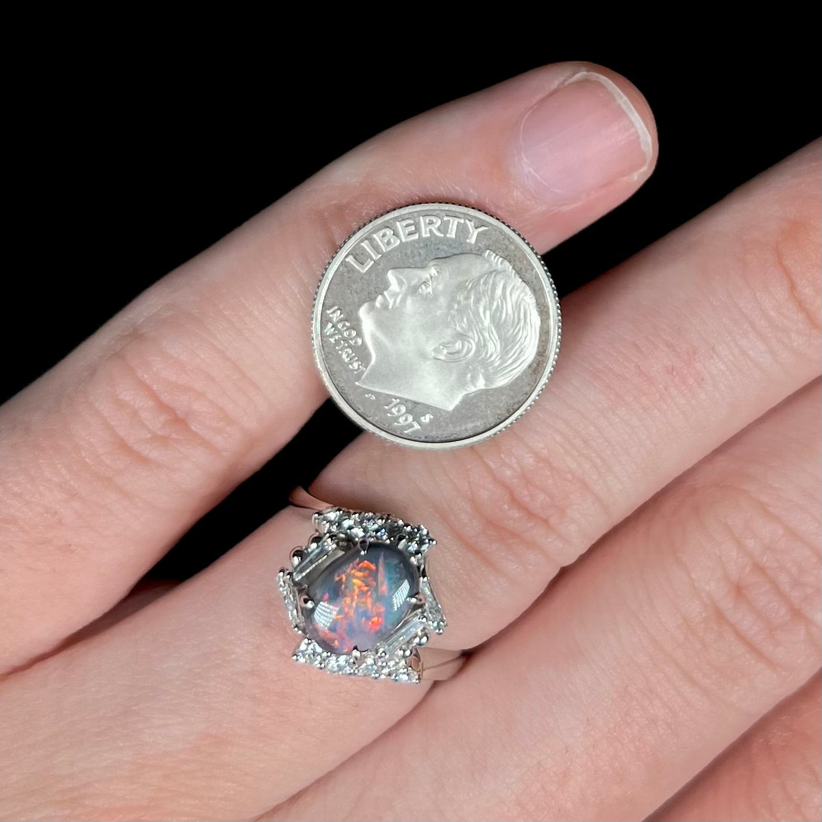 A 1.19ct Australian dark opal ring with geometric diamond halo worn on hand next to US dime coin for scale.