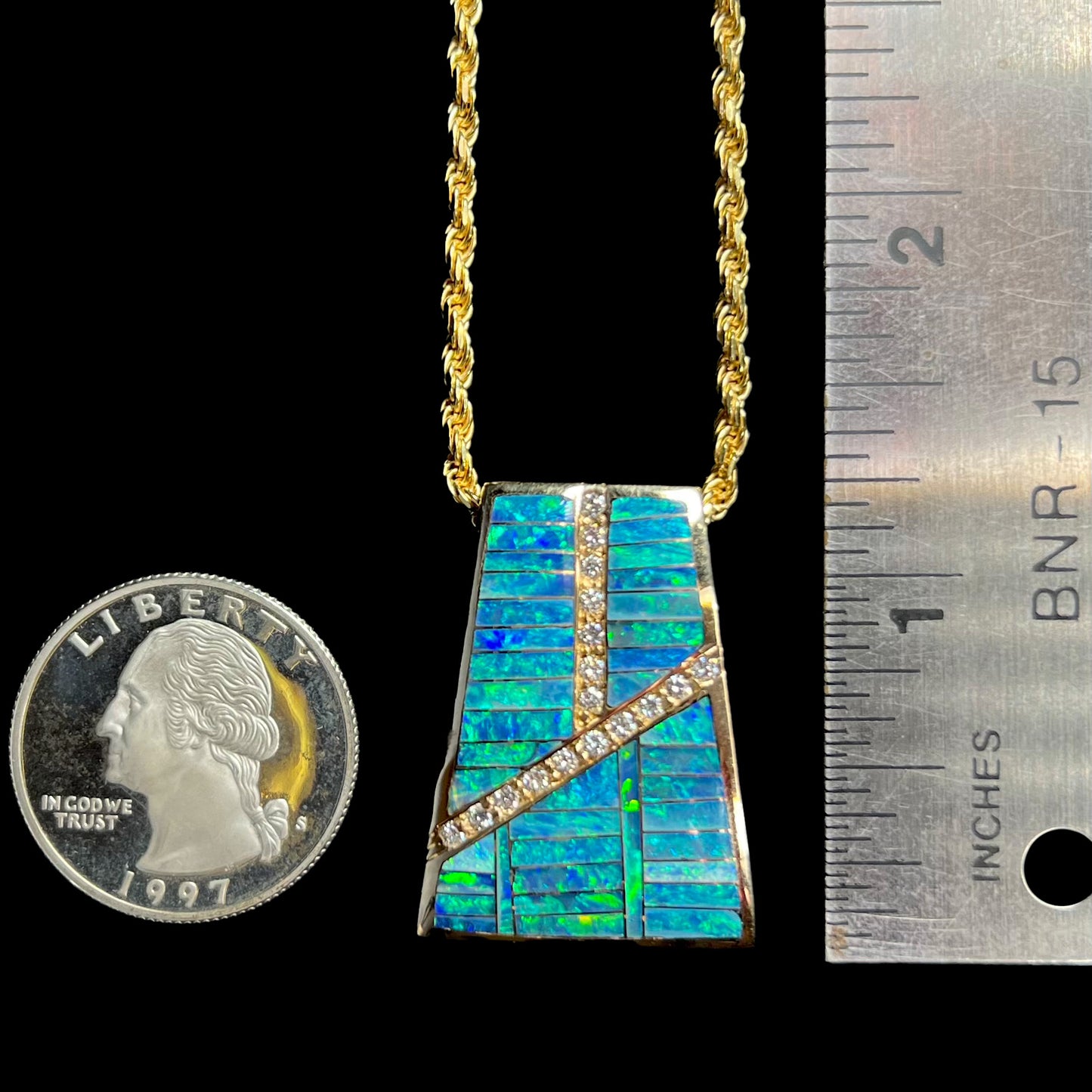 A men's gold opal inlay pendant shown next to a quarter coin and ruler.  The pendant measures 1.25 inches in length.