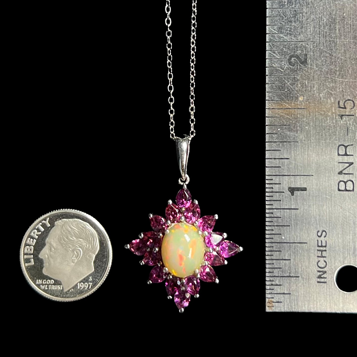 A 1.25 inch long sterling silver Ethiopian opal and rhodolite garnet spray necklace next to a dime coin and ruler.