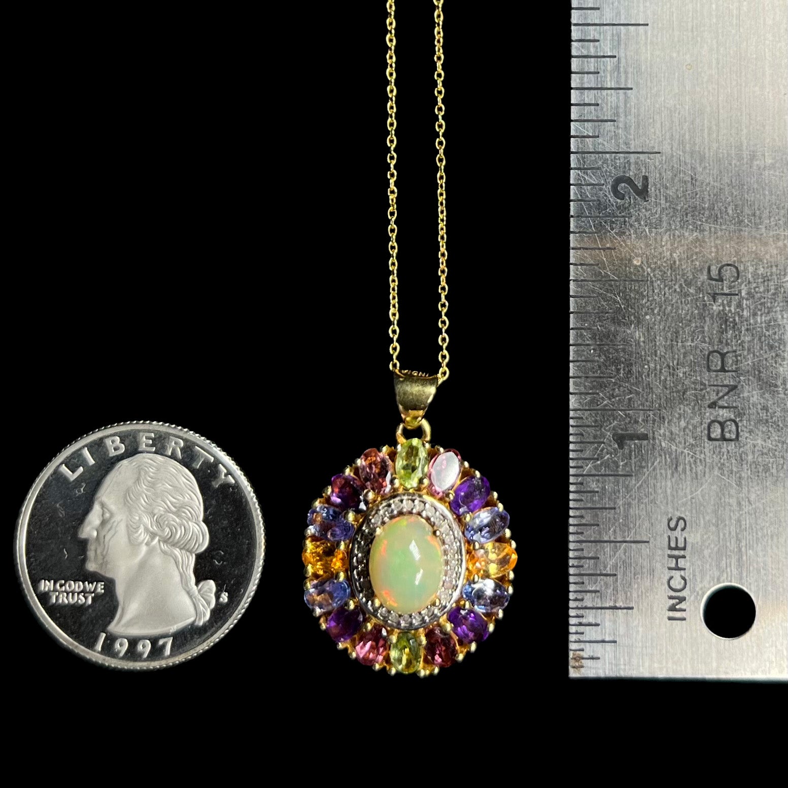 A 1.25 inch long gold plated Ethiopian opal and multi-gemstone halo pendant next to a quarter coin and ruler.