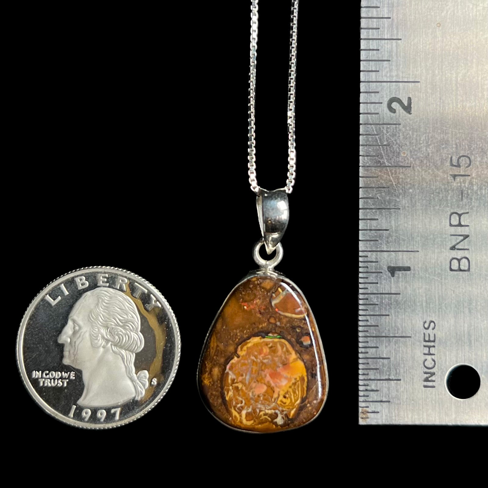 A 1.33 inch long Yowah nut boulder opal pendant in sterling silver, shown next to a ruler and 1997 S US quarter coin.