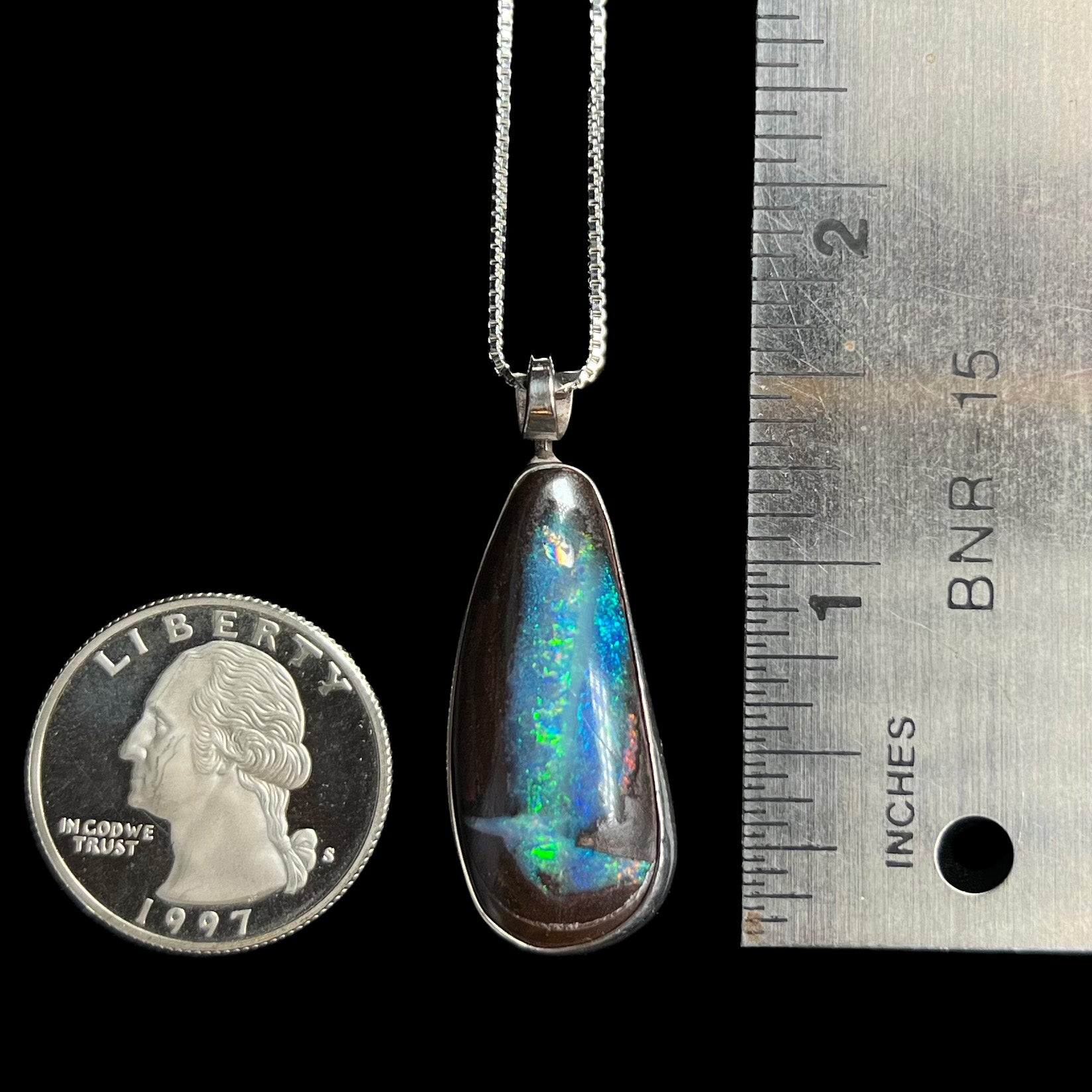 A 1.5 inch-long sterling silver boulder opal pendant with blue-green color play, shown next to a ruler and quarter.
