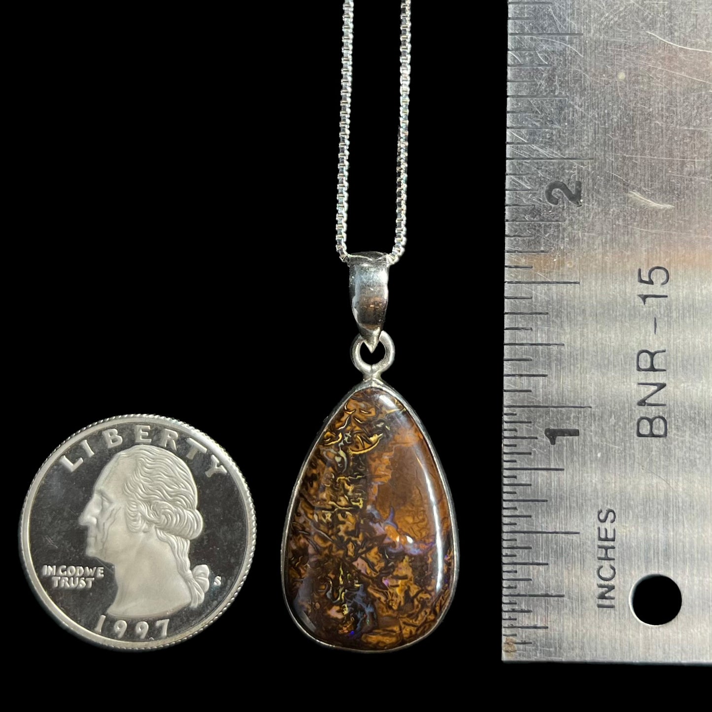 A 1.5 inch-long pear shaped Koroit boulder opal pendant in sterling silver shown next to a ruler and quarter coin.