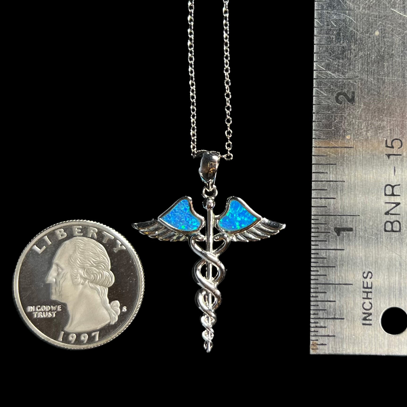 A 1.5 inch long silver plated medical caduceus pendant with lab-created opal inlay next to a quarter and ruler.