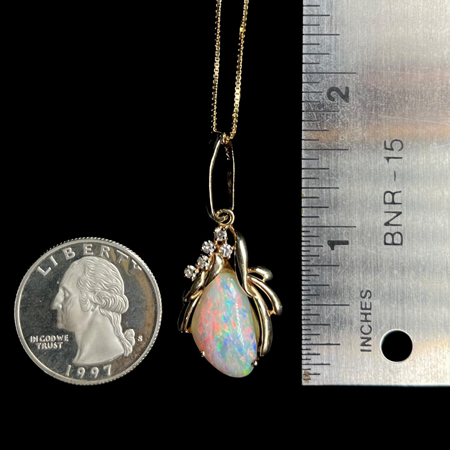 A 1.5 inch-long Modernist-style gold opal and diamond pendant shown next to a US quarter coin and ruler for scale.  The pendant's bail is long.