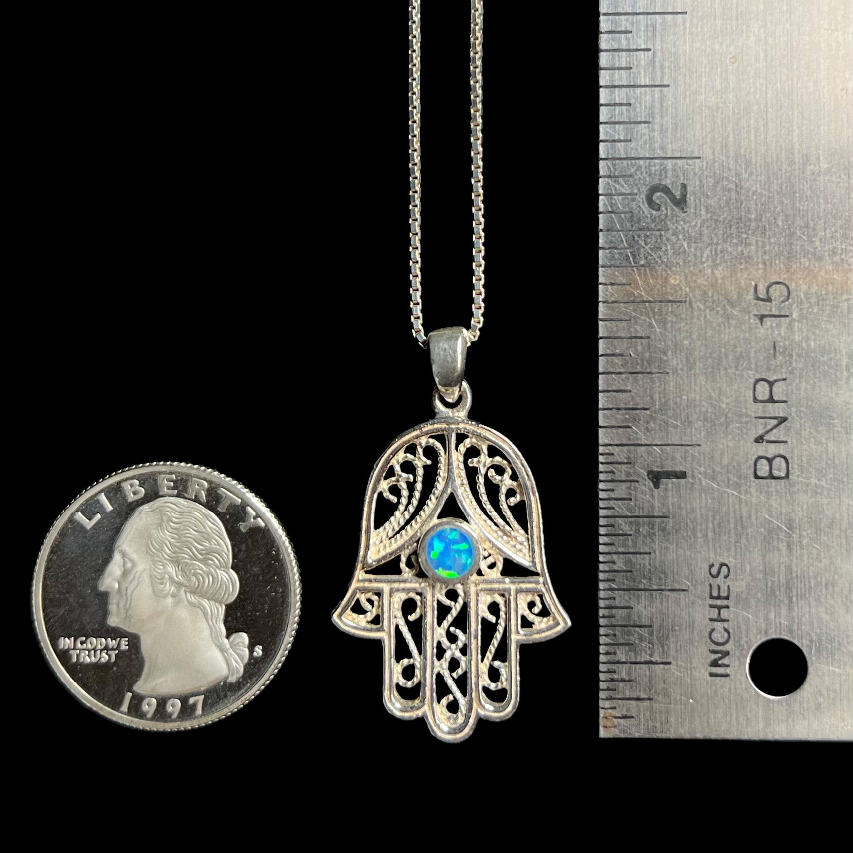 A 1.5 inch long sterling silver hamsa hand necklace set with a round lab-created opal next to a ruler and quarter.