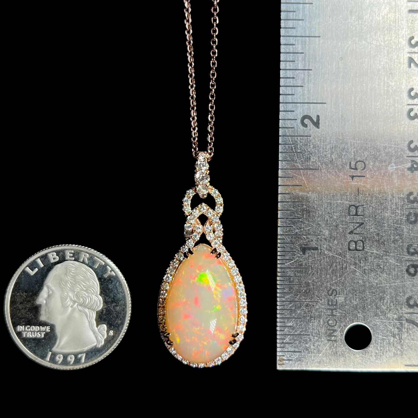 A 1.66 inch-long pear shaped Ethiopian opal and diamond halo necklace shown next to a quarter coin and ruler against a black background.
