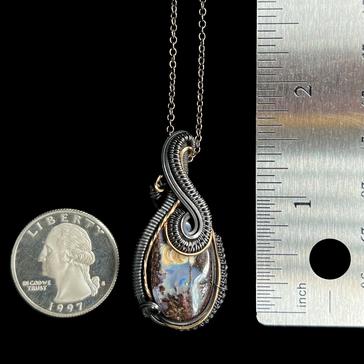 A 1.7 inch-long unisex wire wrapped boulder opal solitaire pendant, shown on a darkened chain between a US quarter coin and ruler for scale.