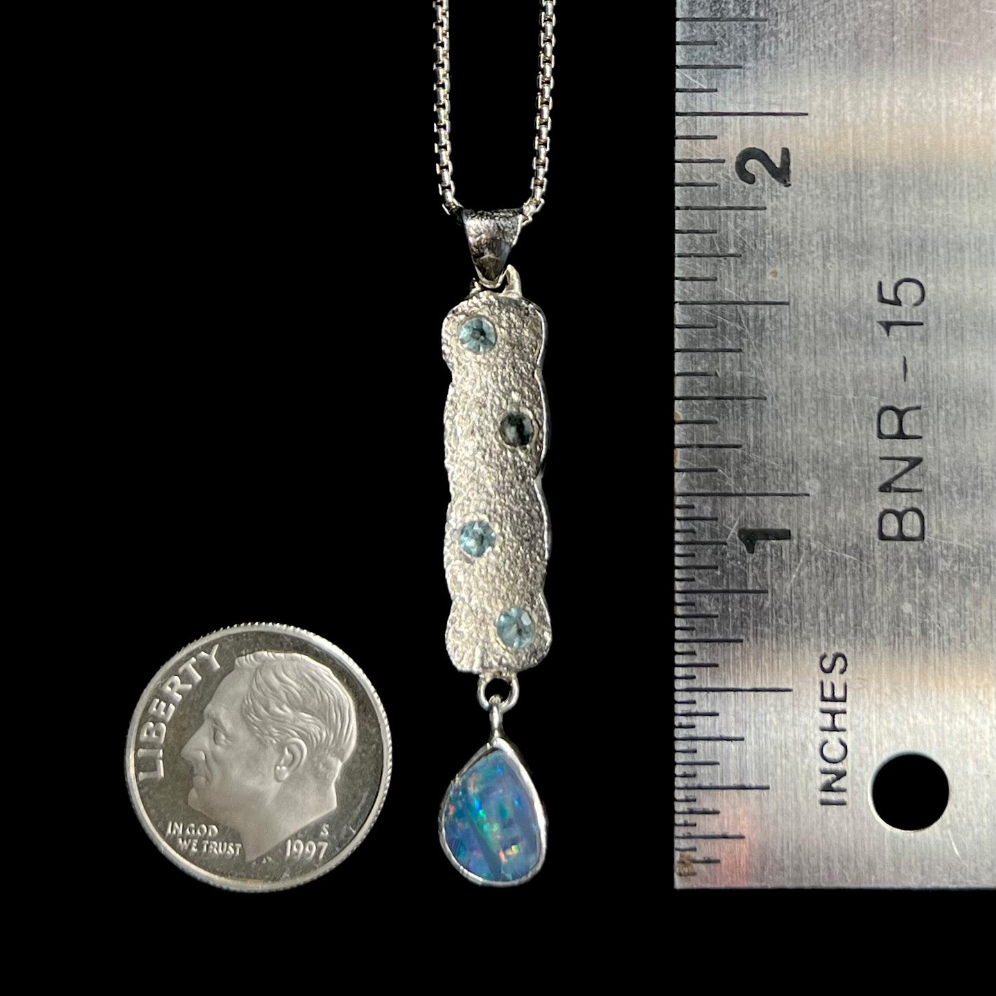 A 1.75 inch-long sterling silver dangling opal doublet drop pendant shown next to a ruler and US dime coin for scale.