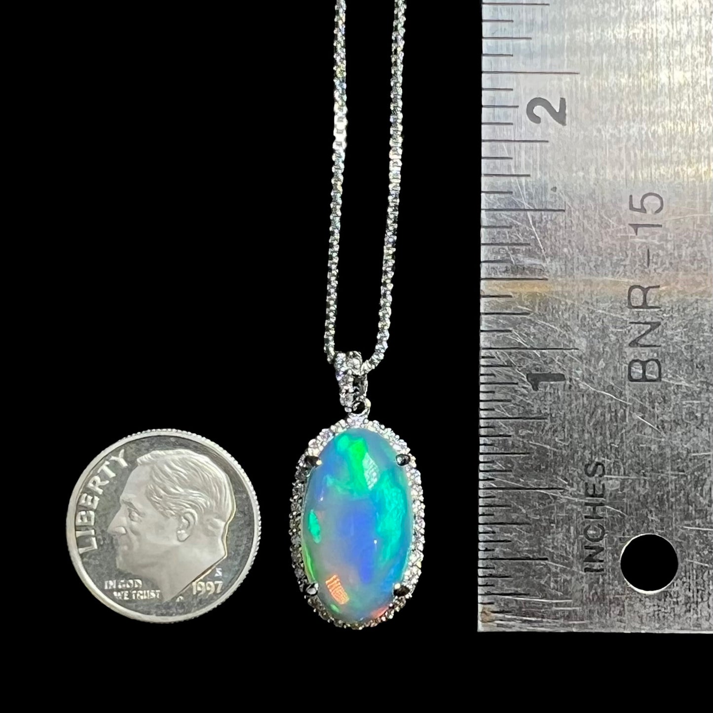 A 1 inch-long platinum Ethiopian opal and diamond halo pendant shown between a dime coin and ruler on a black background.