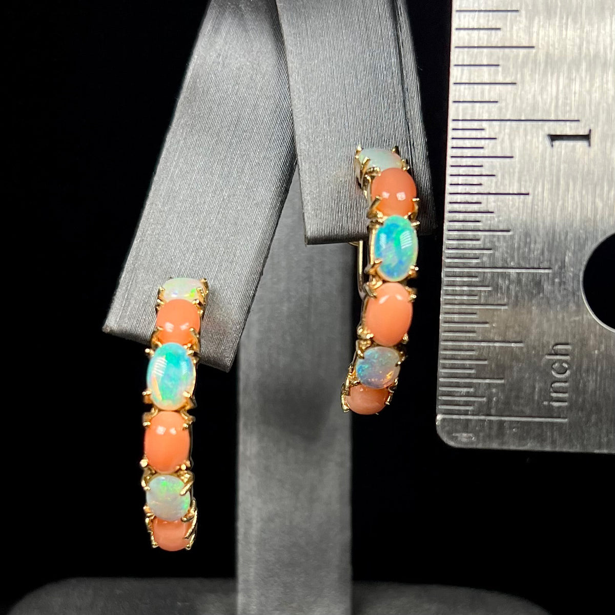 Pair of large opal and coral semi-hoop earrings in 14k gold that measure 1 inch long, shown on earring display next to ruler