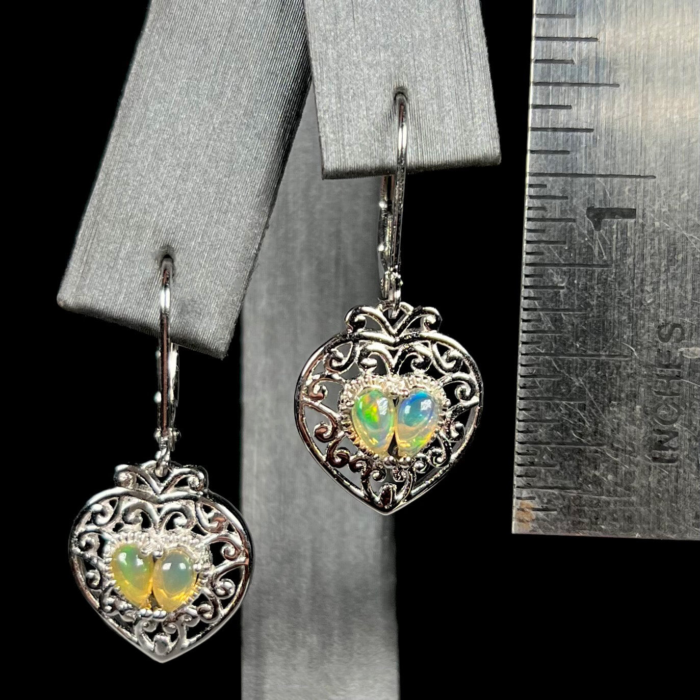 A pair of dangling sterling silver filigree heart earrings, each set with two Ethiopian opals, shown against a ruler.