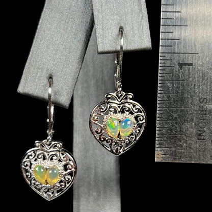 A pair of dangling sterling silver filigree heart earrings, each set with two Ethiopian opals, shown against a ruler.