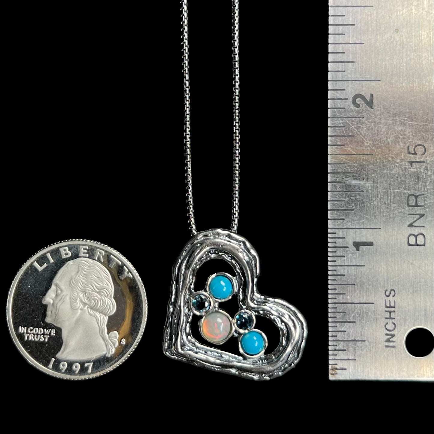 A 1 inch long silver heart necklace set with opal, turquoise, and blue topaz, shown next to quarter coin and ruler.