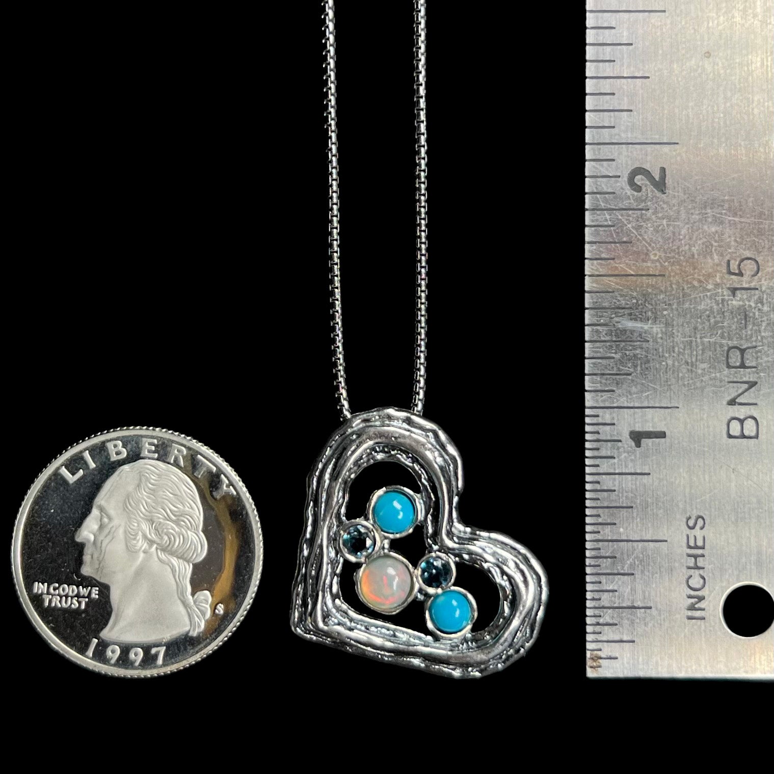 A 1 inch long silver heart necklace set with opal, turquoise, and blue topaz, shown next to quarter coin and ruler.