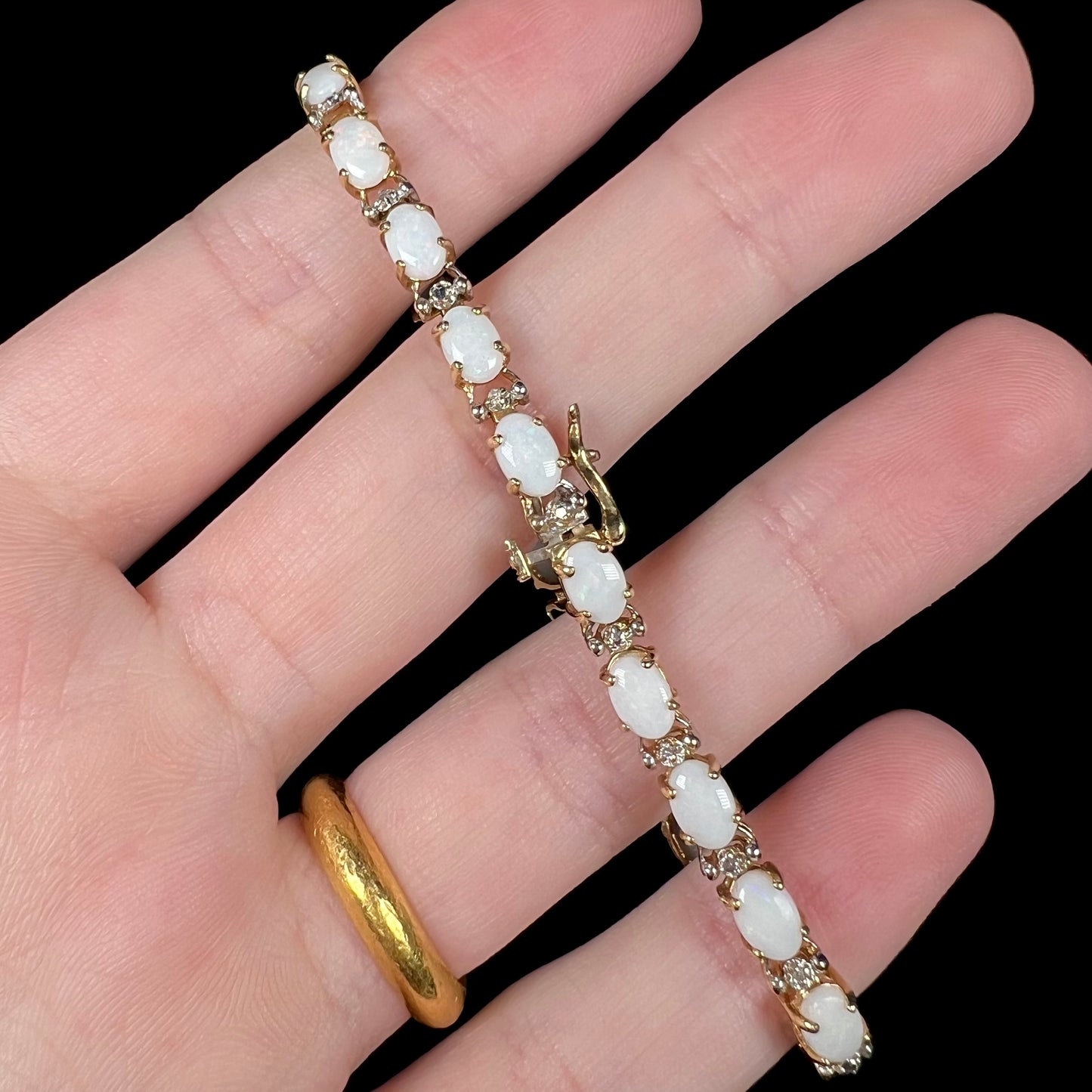 Back side of a 10k gold opal and diamond tennis bracelet shown held in a hand, its box clasp with a single safety latch in focus.