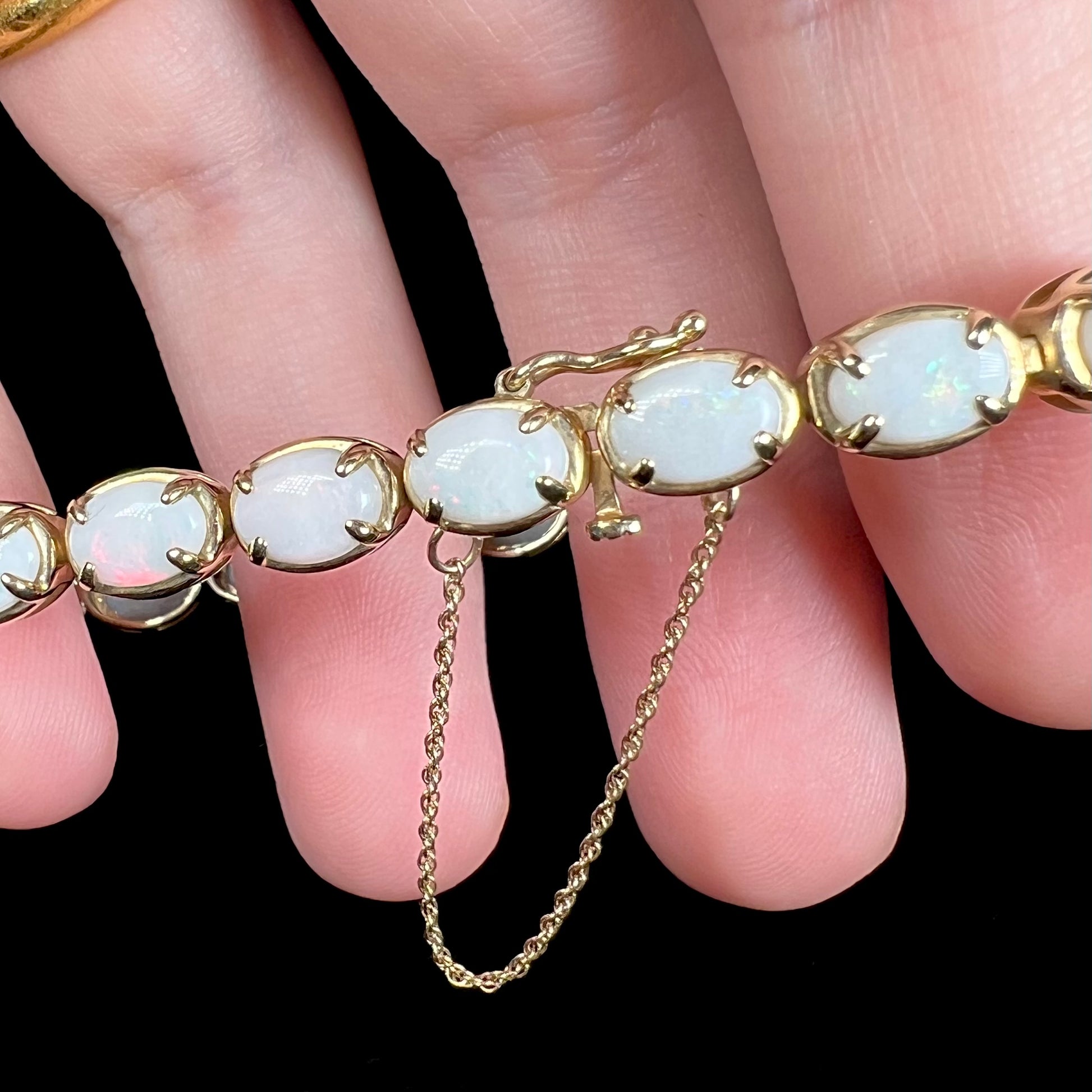 Close-up of 10k gold safety chain and latch on box clasp of an opal tennis bracelet, held in a hand.
