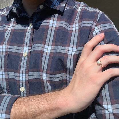 A close-up of a person wearing a navy, brown, and gray plaid button-down shirt, with their right hand resting on their left shoulder. The person is wearing a 10k yellow gold ring with an Australian opal on their ring finger.