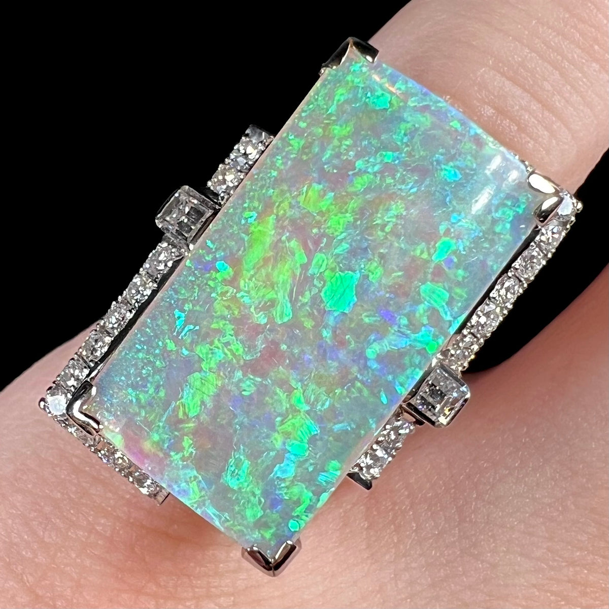 Close-up of rectangle cut 11.46ct Australian light opal and diamond cocktail ring, worn on pinky finger.  The opal shines with patches of green, blue, and teal play-of-color under LED studio light.