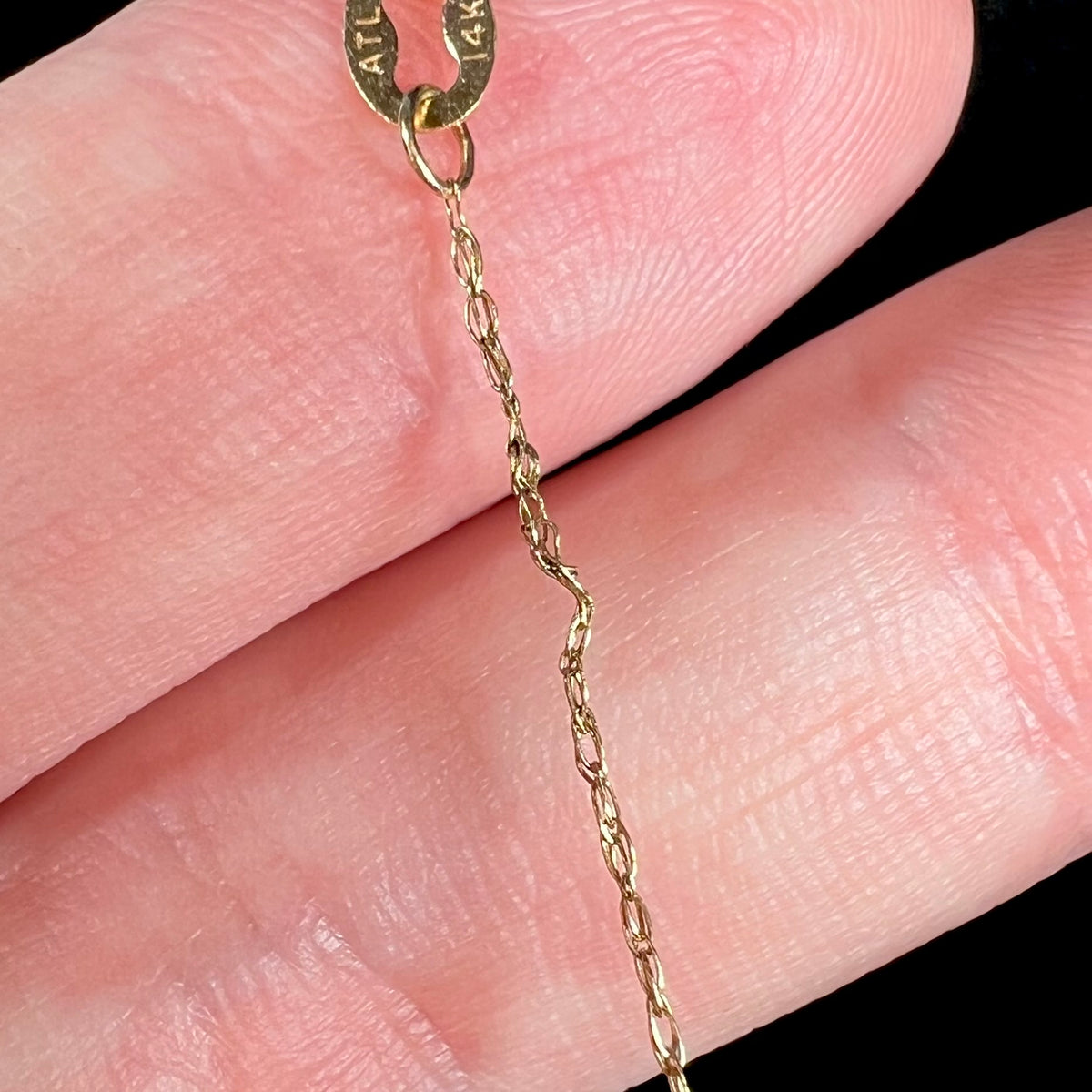 Close-up of a solder repair point on a dainty 14k yellow gold chain; the chain's tag is seen, reading the hallmark stamps "ATL" and "14K"