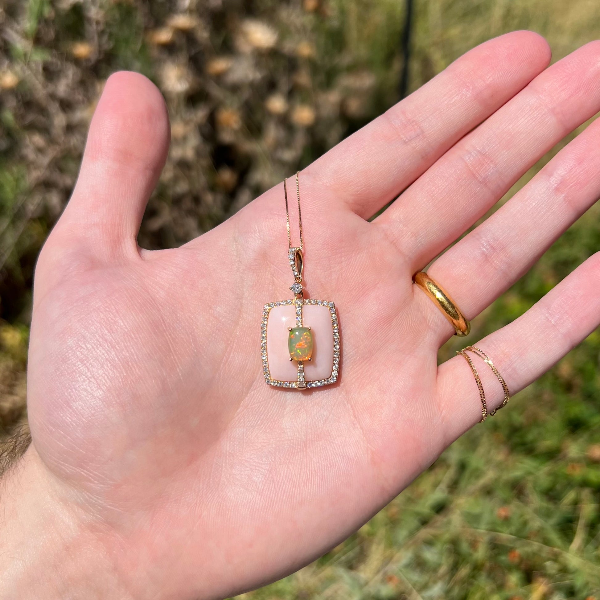 Hand holding a cushion cut fire opal, Peruvian pink opal, and diamond halo necklace with a chain in 14k gold in the sunlight.