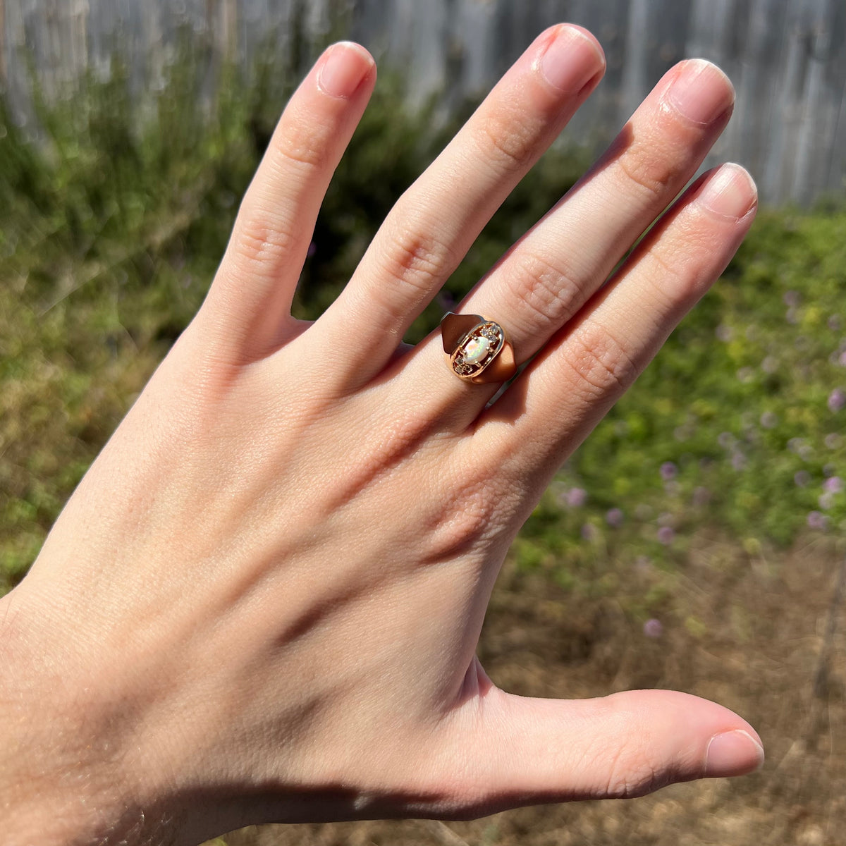 Sunlit hand wearing a 14k gold Mid-Century style opal and diamond ring with domed design against blurred green and brown outdoor background