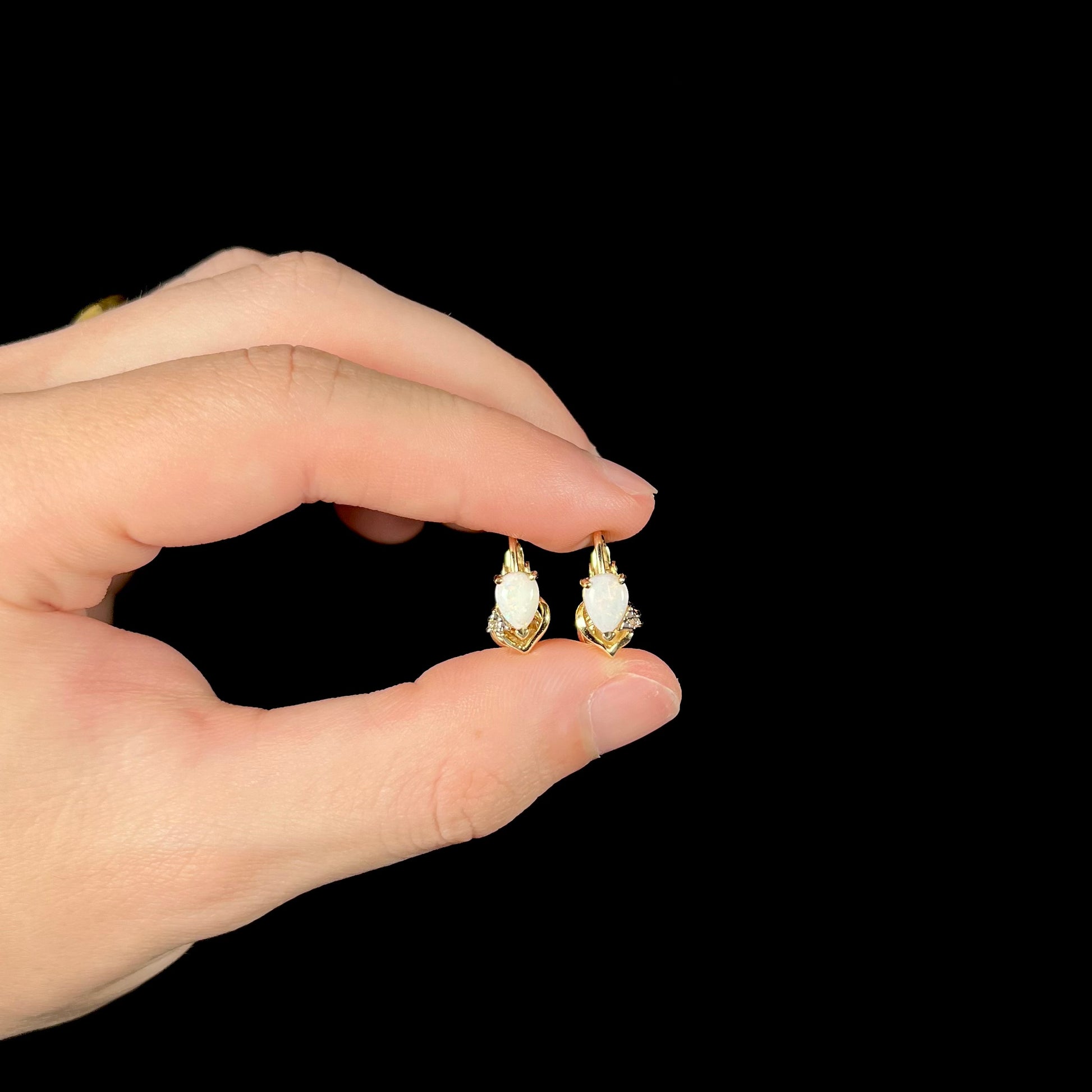 A pair of 14k gold opal and diamond clip-on earrings held in a hand, showing their size and elegant design against a black background.