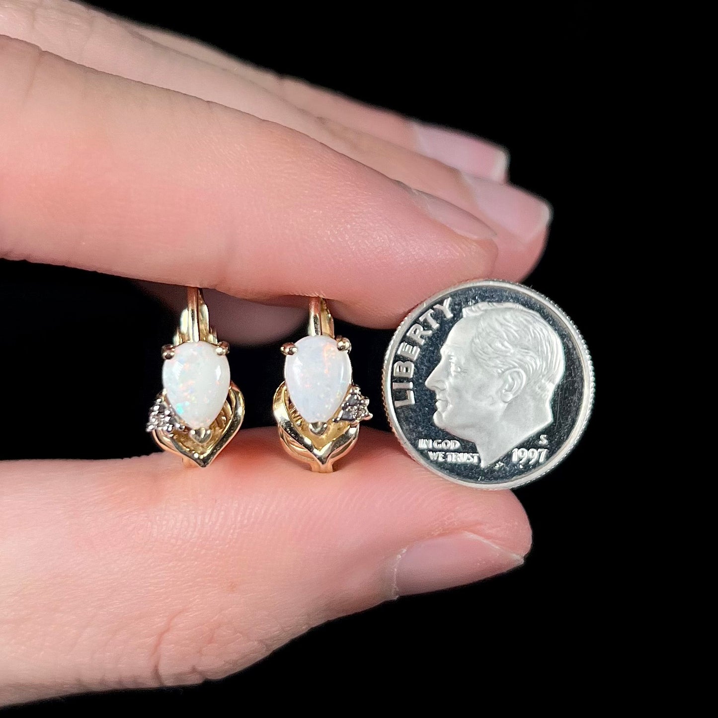 A macro close-up of 14k gold opal and diamond clip-on earrings held between fingers with a dime for scale, highlighting their petite size and natural opal fire.