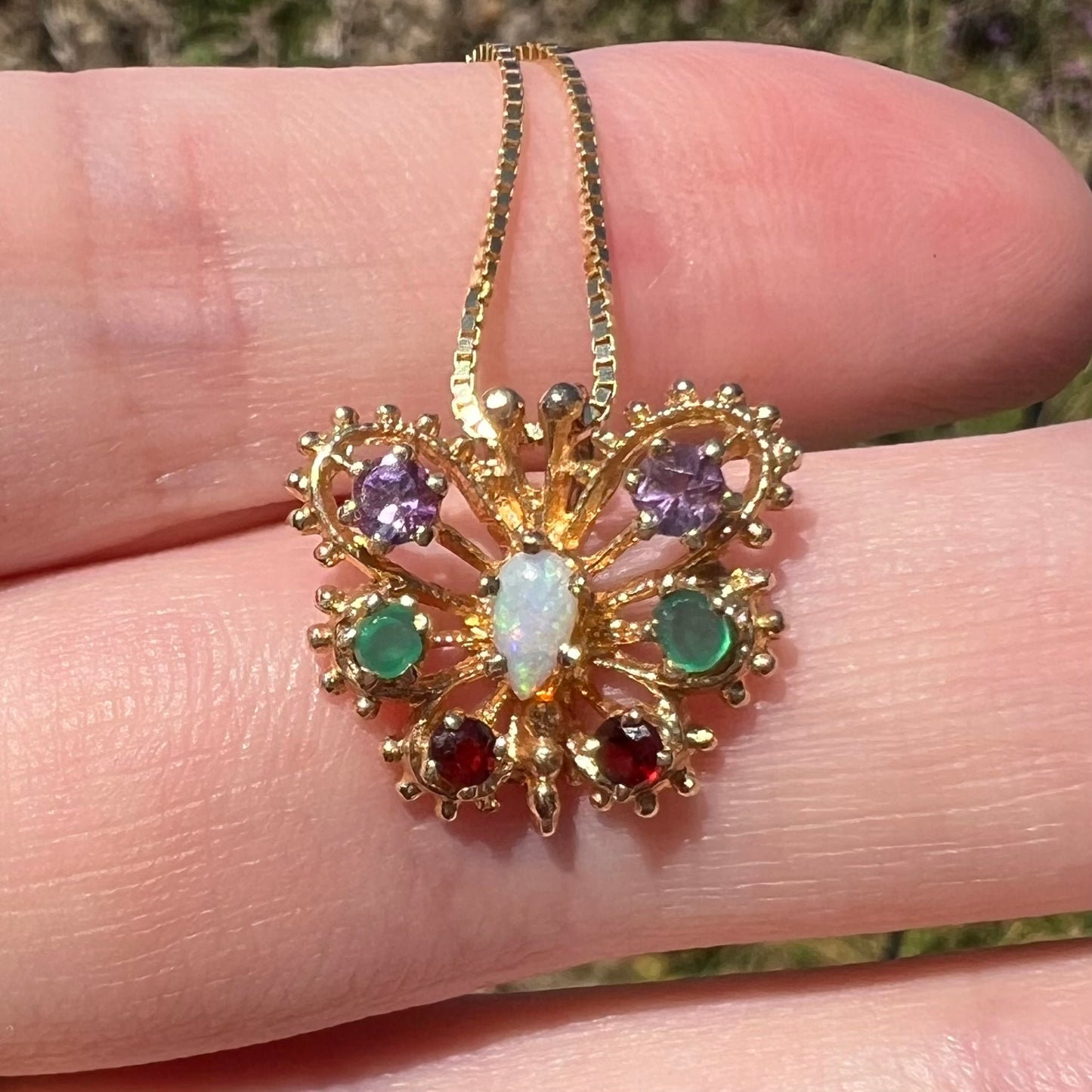 Close-up of a vintage 14k yellow gold opal, amethyst, emerald, and garnet gemstone butterfly pendant in natural light, resting on top of fingers.