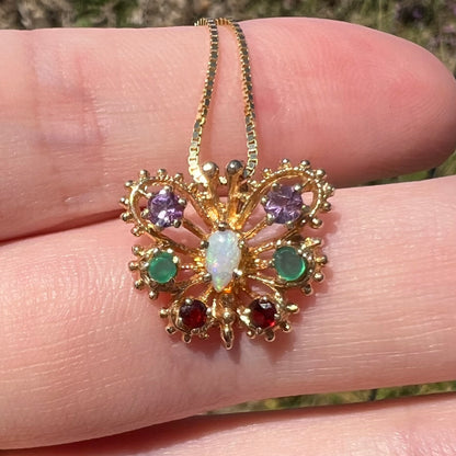 Close-up of a vintage 14k yellow gold opal, amethyst, emerald, and garnet gemstone butterfly pendant in natural light, resting on top of fingers.