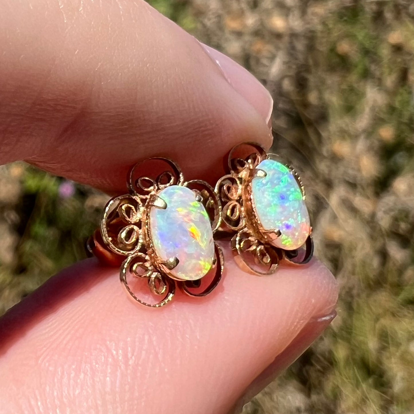 Angled close-up of 14k gold opal stud earrings with filigree flower design in sunlight, held between fingers