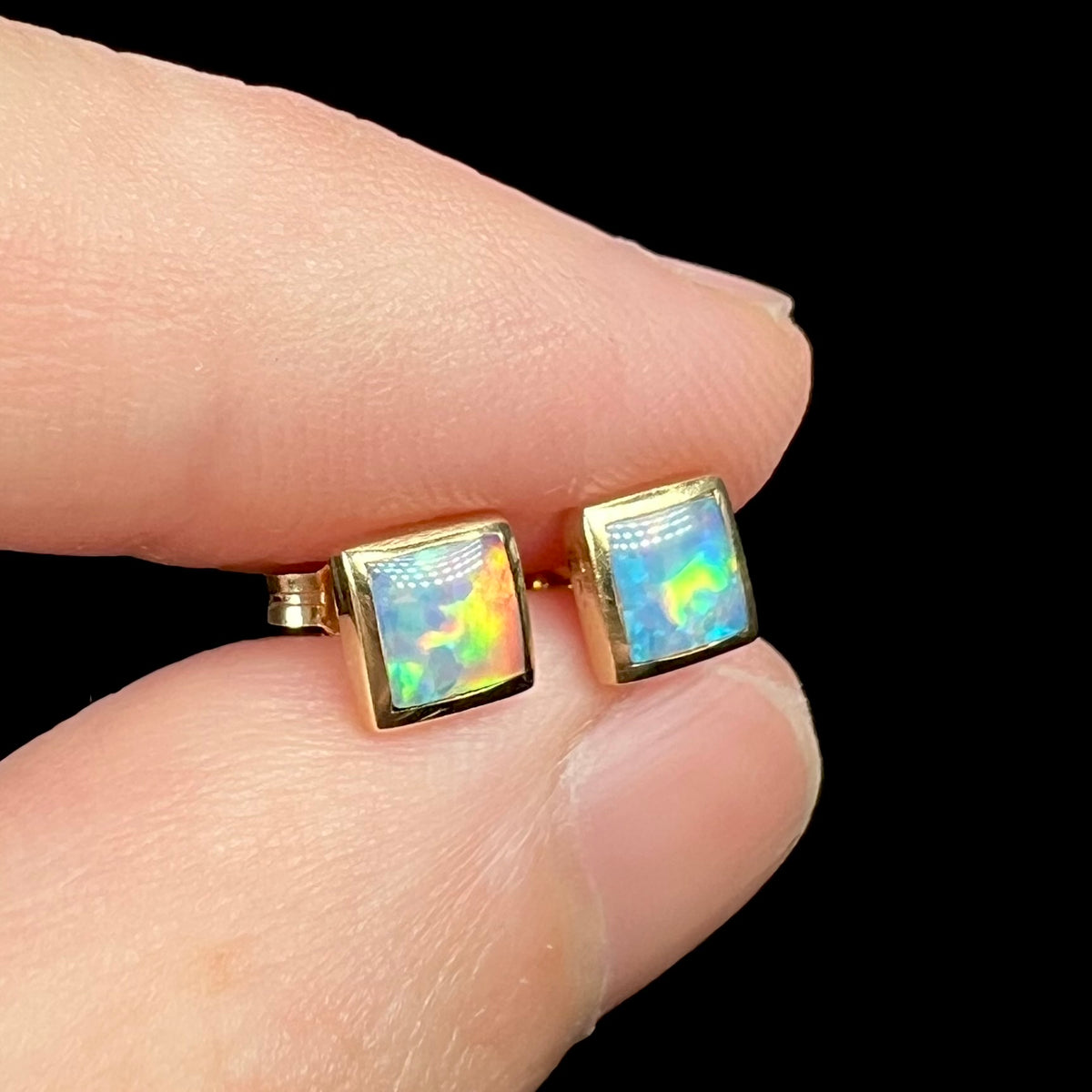 Angled close-up of 14k gold opal stud earrings, held in fingers.  Opals are square shaped and have multicolor flashes