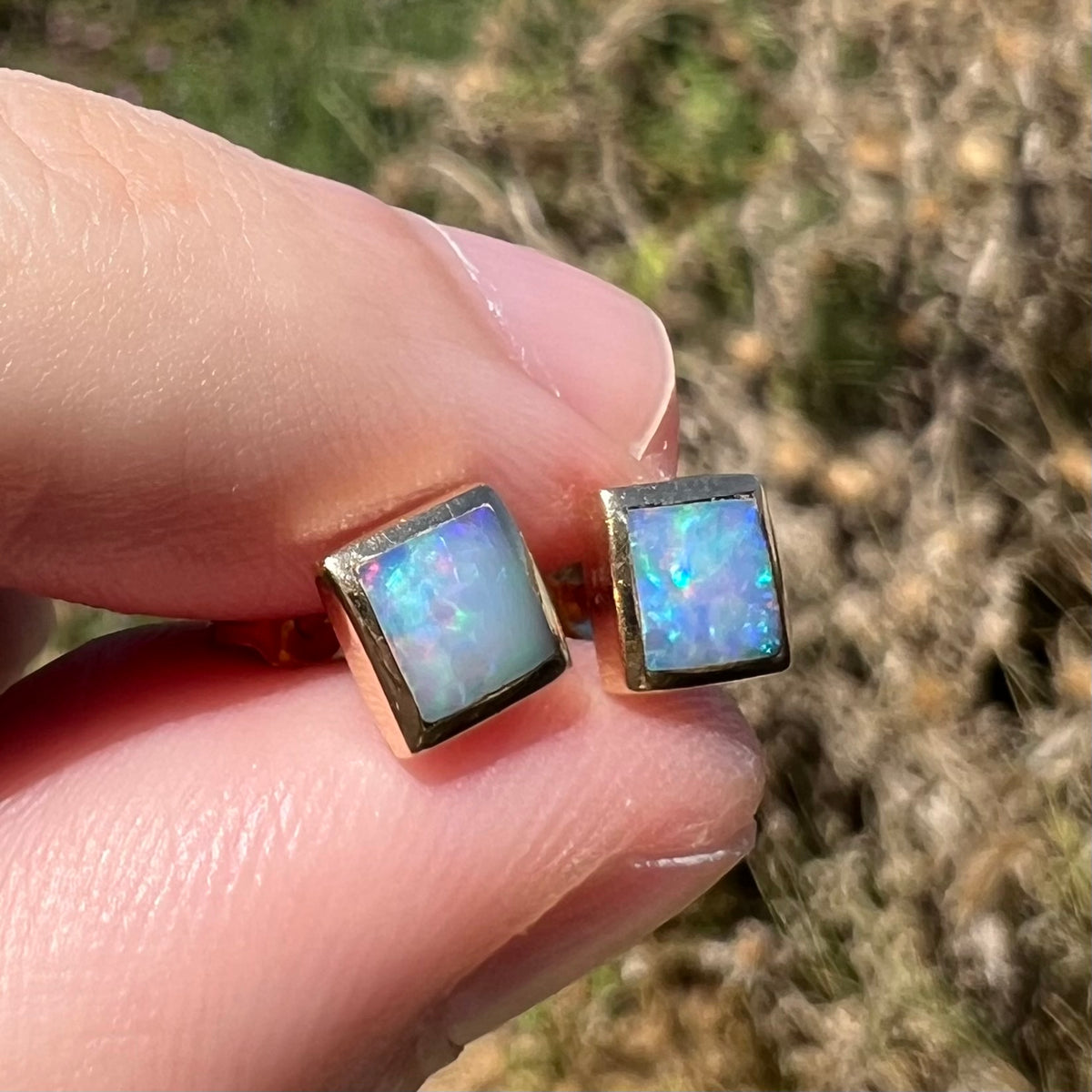 14k gold stud earrings with square shaped opals, held between two fingers in natural light