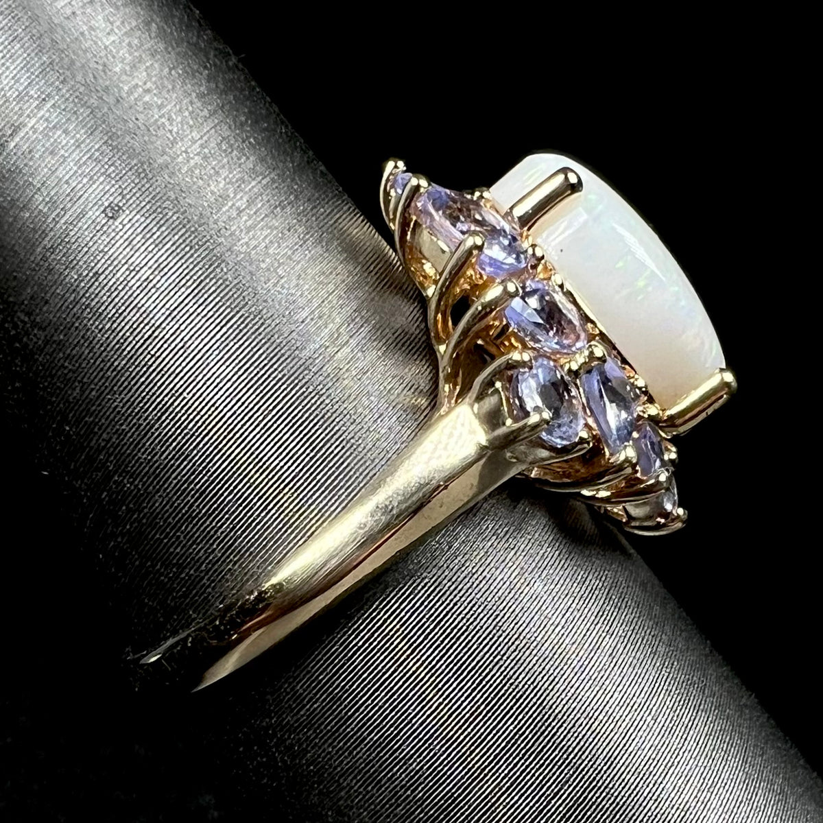 A 14k yellow gold white opal and tanzanite-accented ring shown on a ring display from the side view, the ring's polished shank in focus
