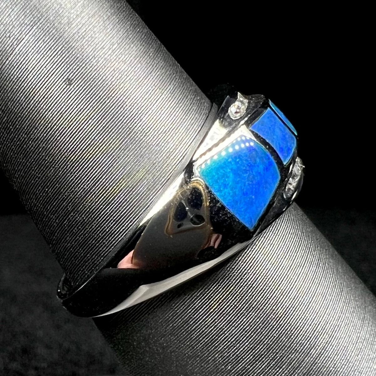 A 14k white gold opal inlay ring shown on ring display from side view, its shank and blue opal side inlay in focus