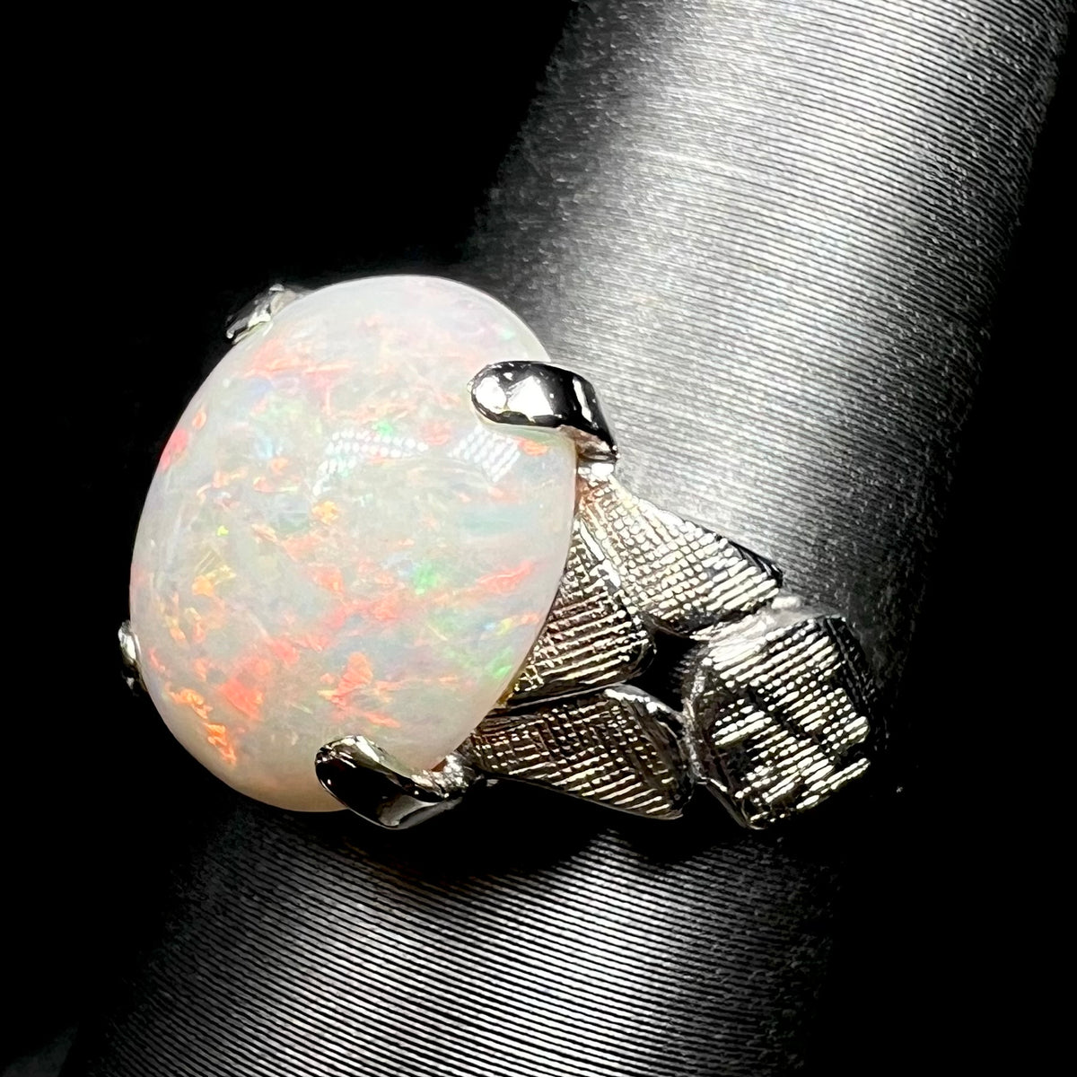 Angled close-up of a 14k white gold natural opal ring with a textured shoulder design, shown on ring display