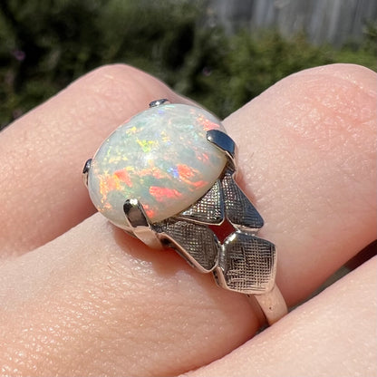 Red and orange colors play in an Australian opal ring, worn on a finger in sunlight; the ring is shown close-up from an angle, its textured 14k white gold shoulders in focus