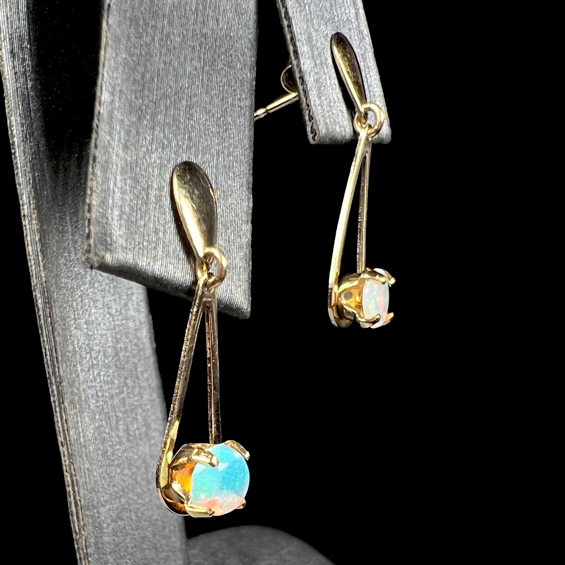 14k yellow gold opal dangle earrings shown from angle on earring display against black background.  Greenish blue opal is in focus.