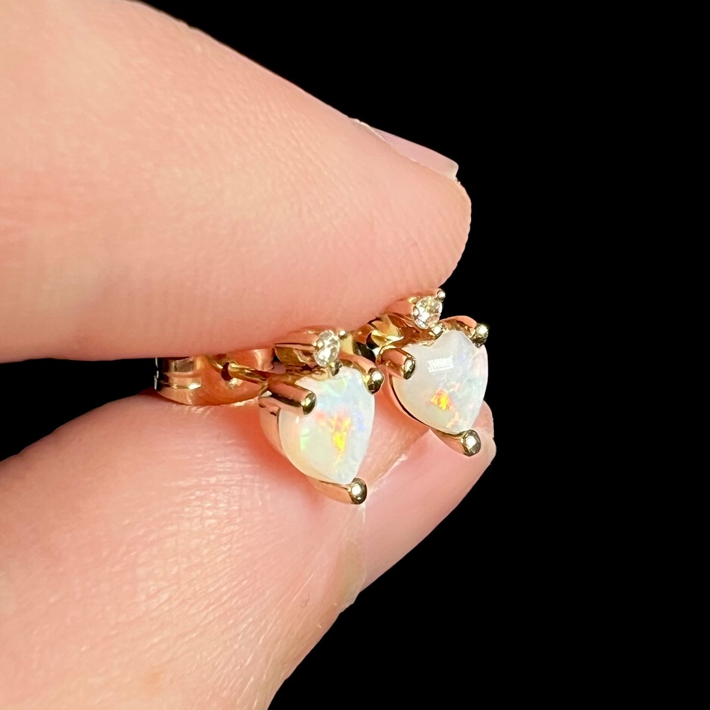 Angled close-up of 14k yellow gold opal heart stud earrings, held between fingers.  The opals have orange and red flashes.